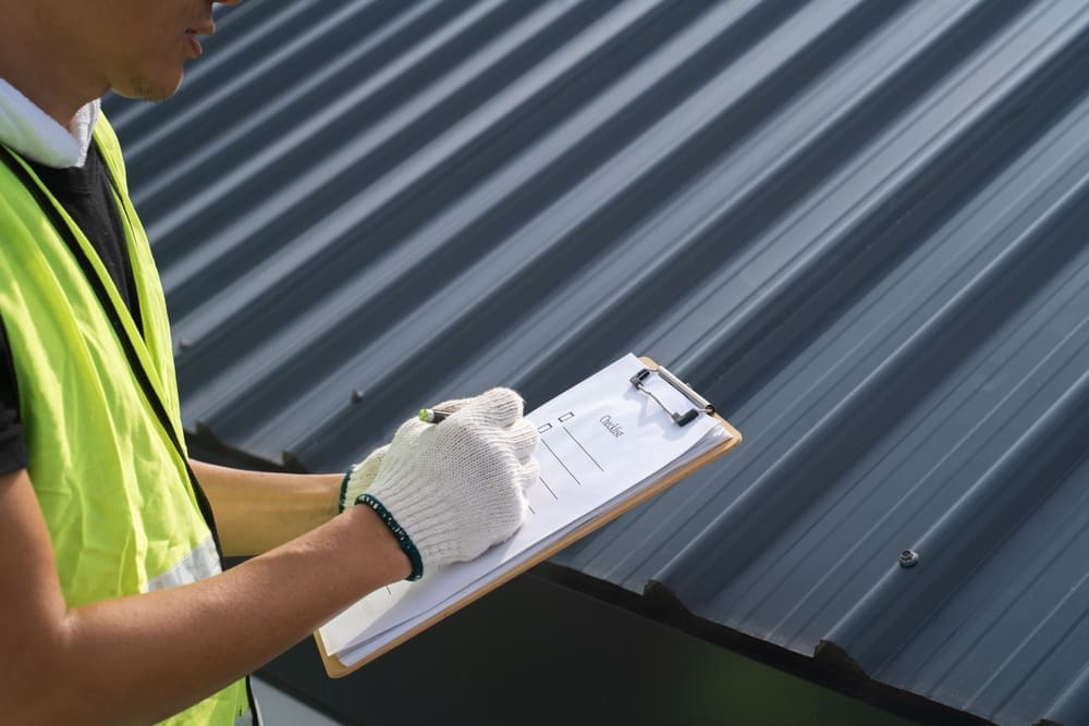 Roofing contractor inspecting a metal roof and recording notes on a clipboard
